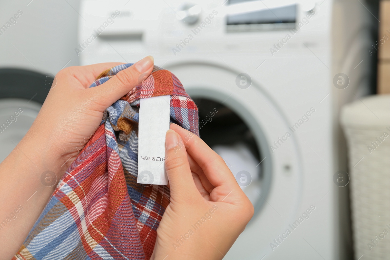 Photo of Woman holding checkered shirt with blank clothing label at home, closeup. Space for text