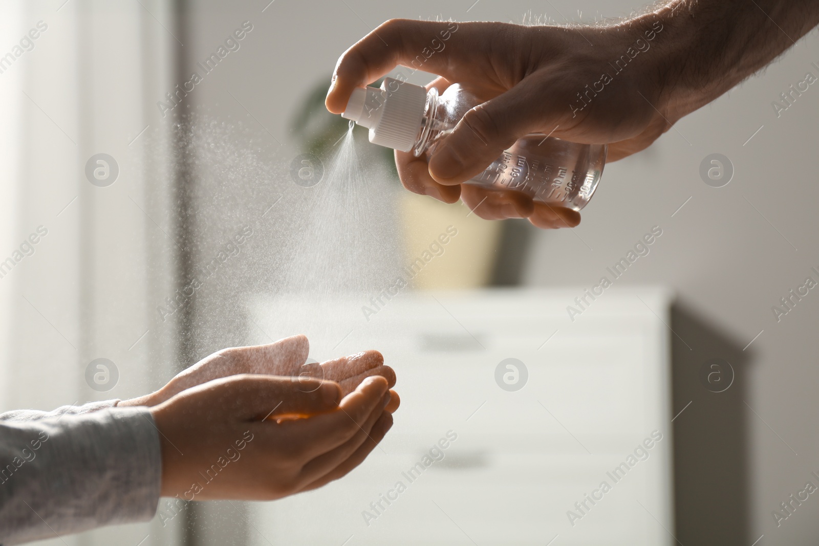 Photo of Man spraying antiseptic onto child's hands indoors, closeup