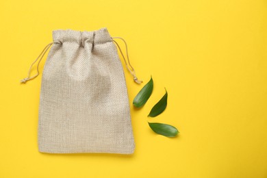 Photo of Burlap bag and green leaves on yellow background, flat lay. Space for text