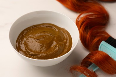 Photo of Bowl of henna cream, brush and red strand on white table, closeup. Natural hair coloring