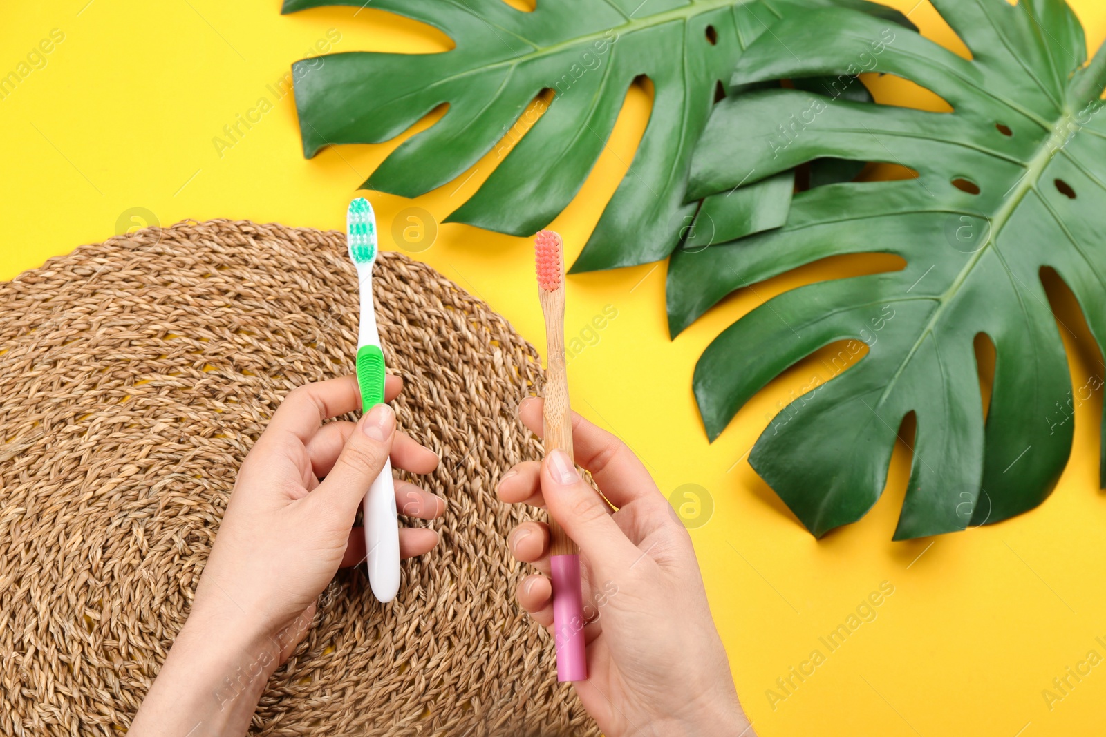 Photo of Woman holding natural bamboo and plastic toothbrushes above yellow background, top view