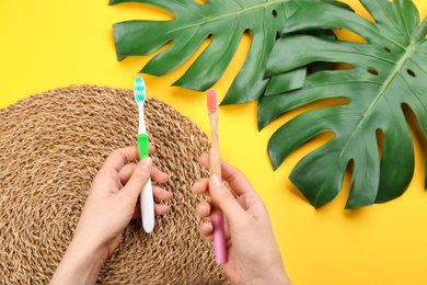 Woman holding natural bamboo and plastic toothbrushes above yellow background, top view