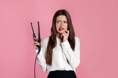 Photo of Upset young woman with flattening iron on light pink background. Hair damage