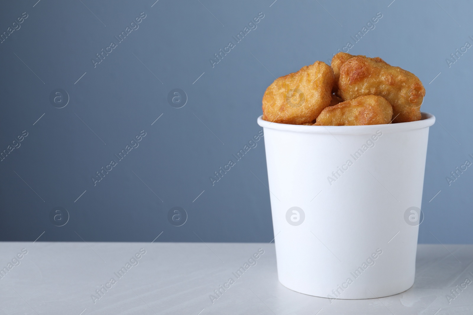 Photo of Bucket with tasty chicken nuggets on light grey table against blue background. Space for text