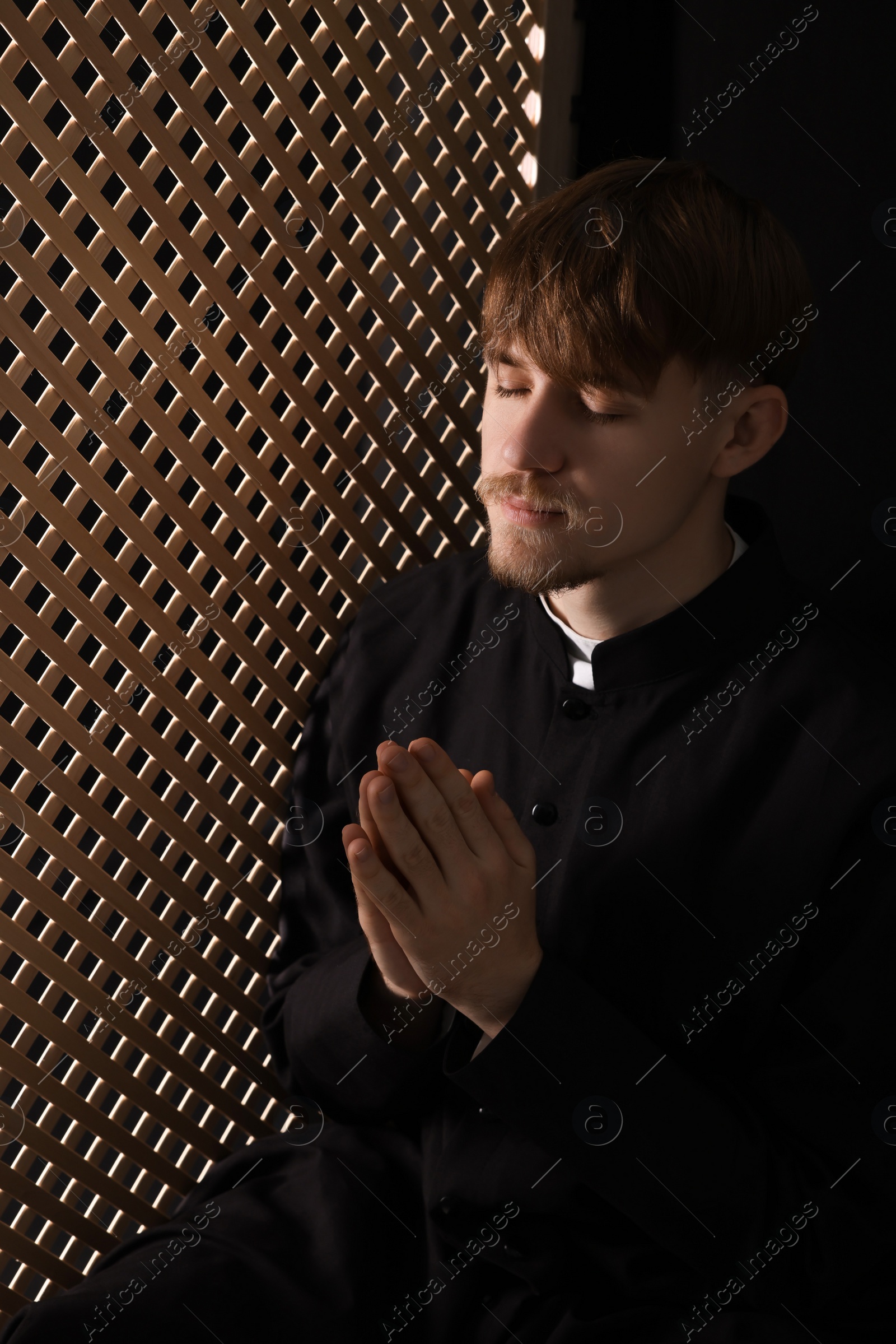 Photo of Catholic priest praying near wooden partition in confessional