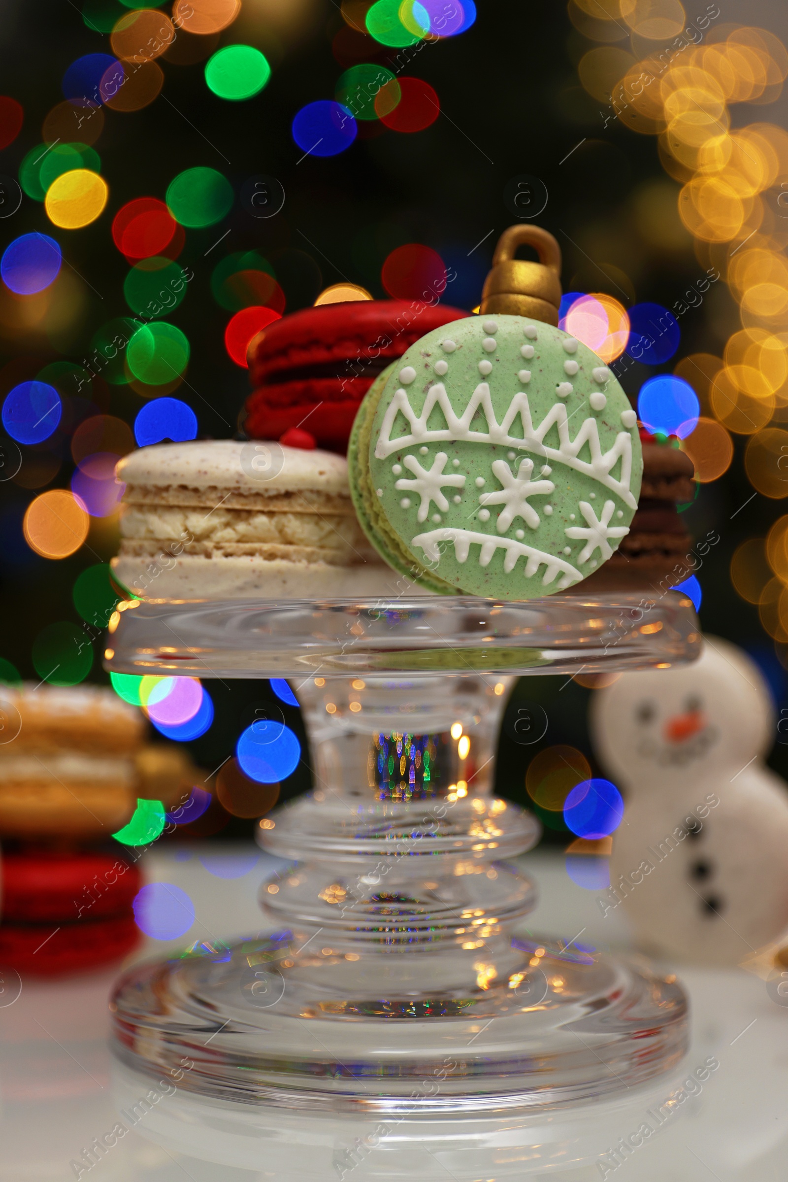 Photo of Beautifully decorated Christmas macarons on white table against blurred festive lights