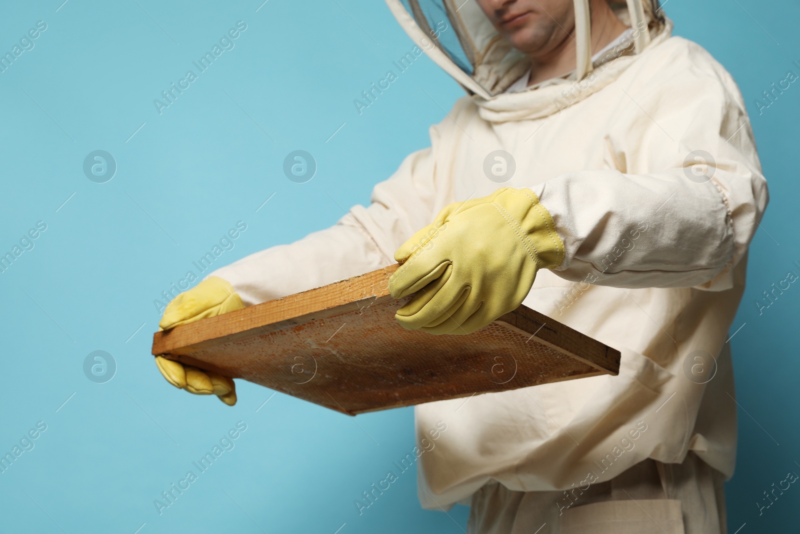 Photo of Beekeeper in uniform holding hive frame with honeycomb on light blue background, closeup