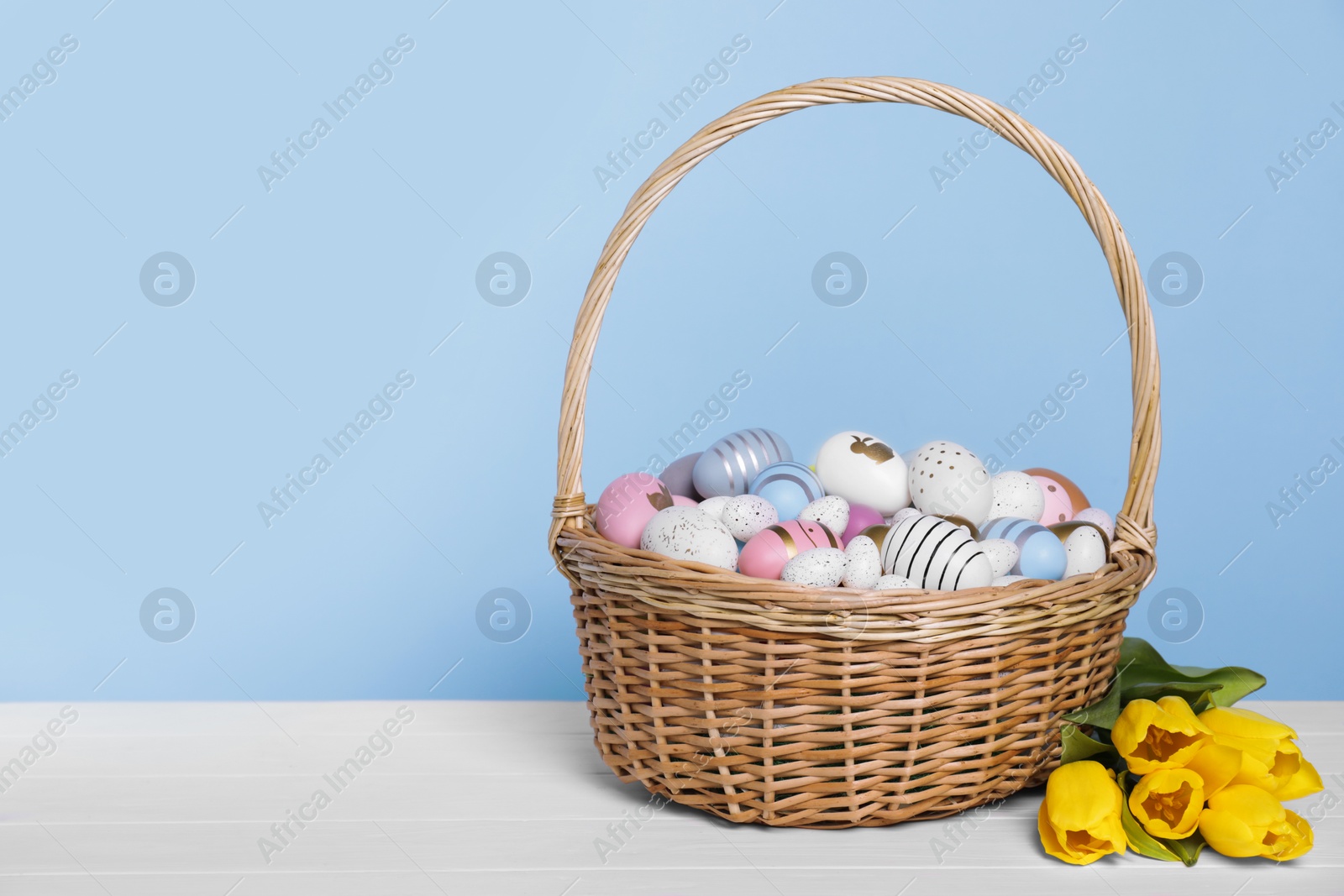 Photo of Wicker basket with festively decorated Easter eggs and beautiful tulips on white wooden table against light blue background. Space for text