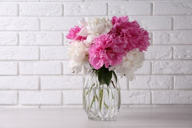 Beautiful peonies in glass vase on white table near brick wall