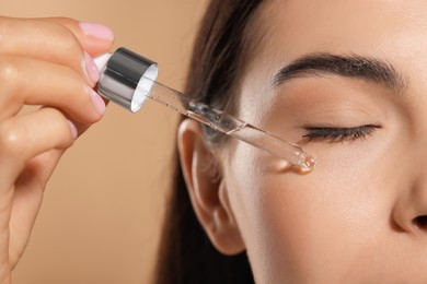 Young woman applying serum onto her face on beige background, closeup