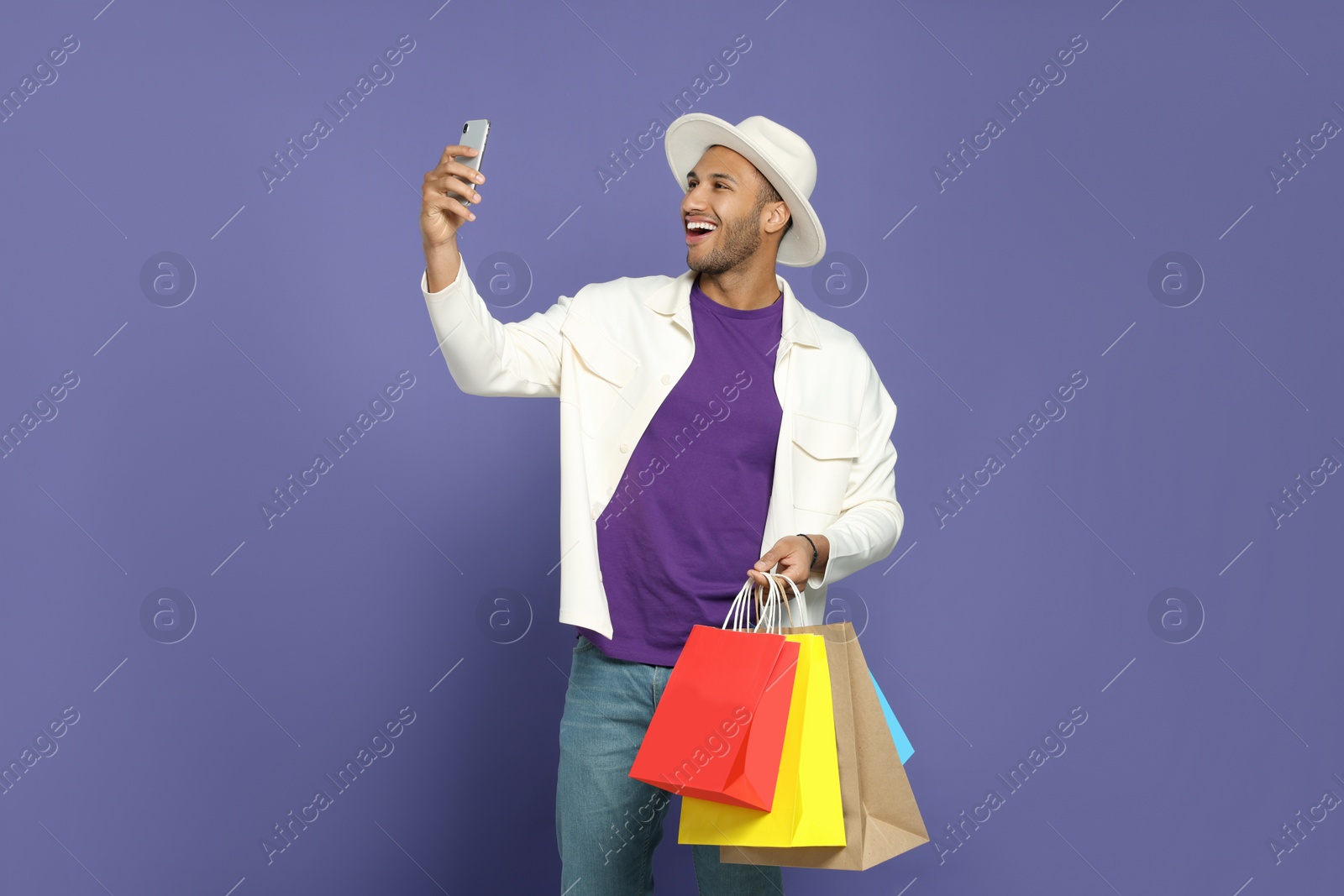 Photo of Happy African American man in hat with shopping bags taking selfie on purple background