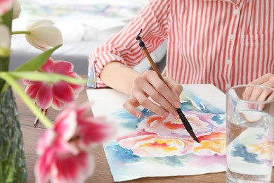 Photo of Woman painting flowers with watercolor at wooden table indoors, closeup. Creative artwork