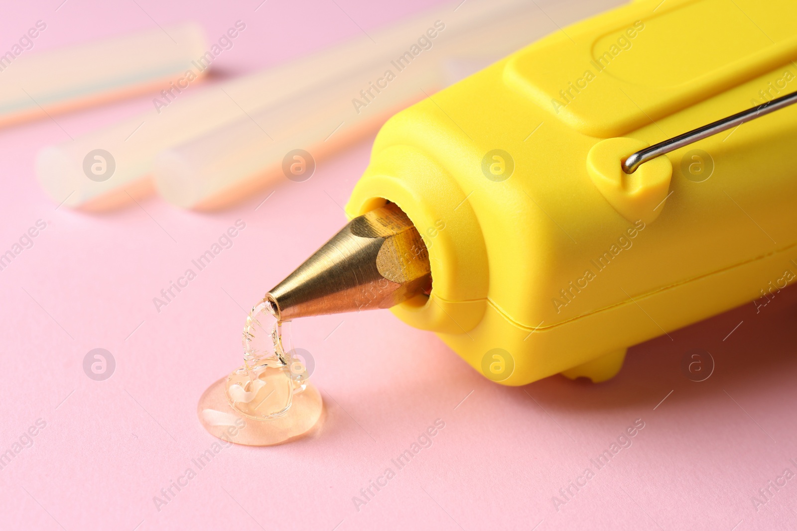 Photo of Melted glue dripping out of hot gun nozzle near sticks on pink background, closeup