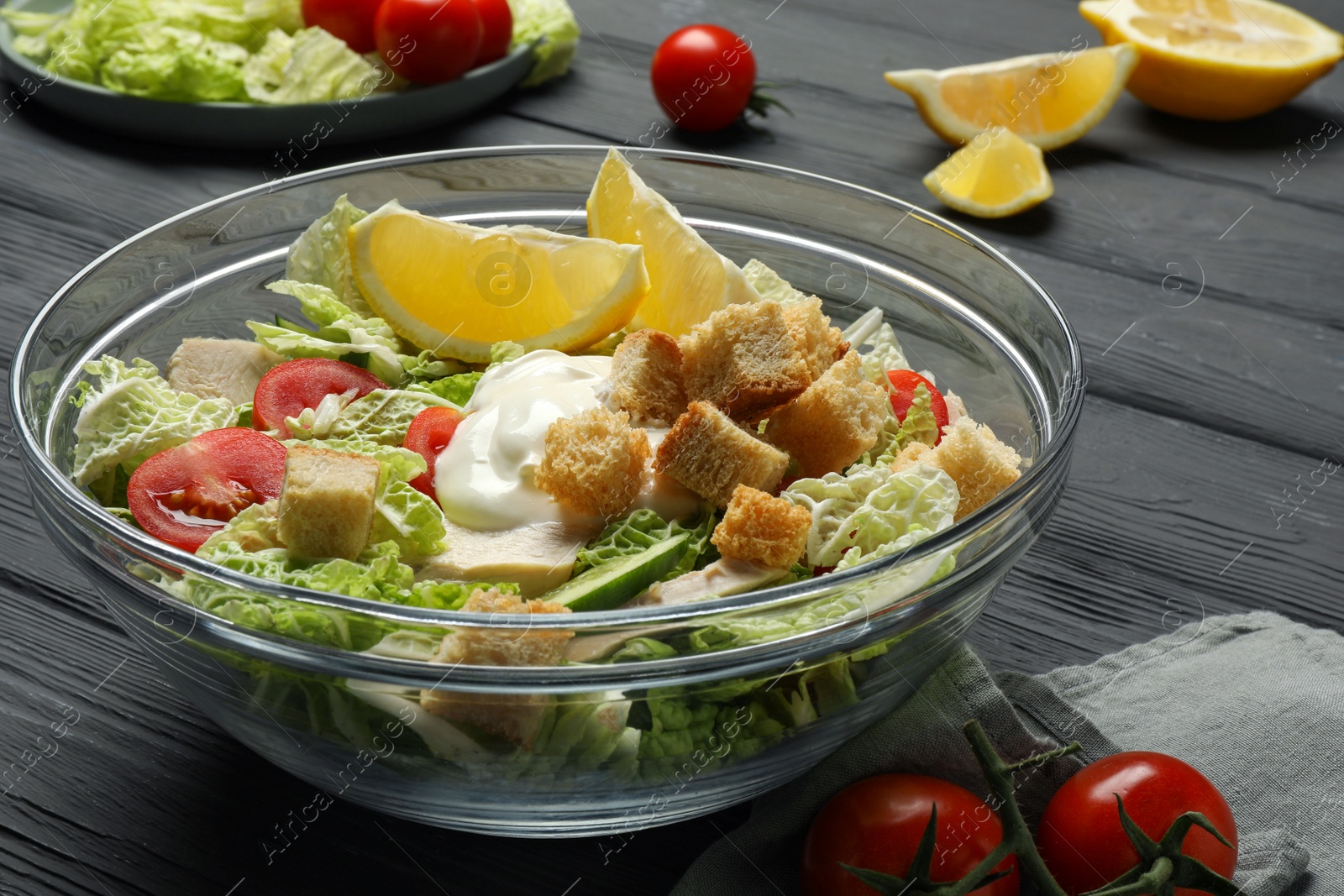 Photo of Bowl of delicious salad with Chinese cabbage, lemon, tomatoes and bread croutons on black wooden table, closeup