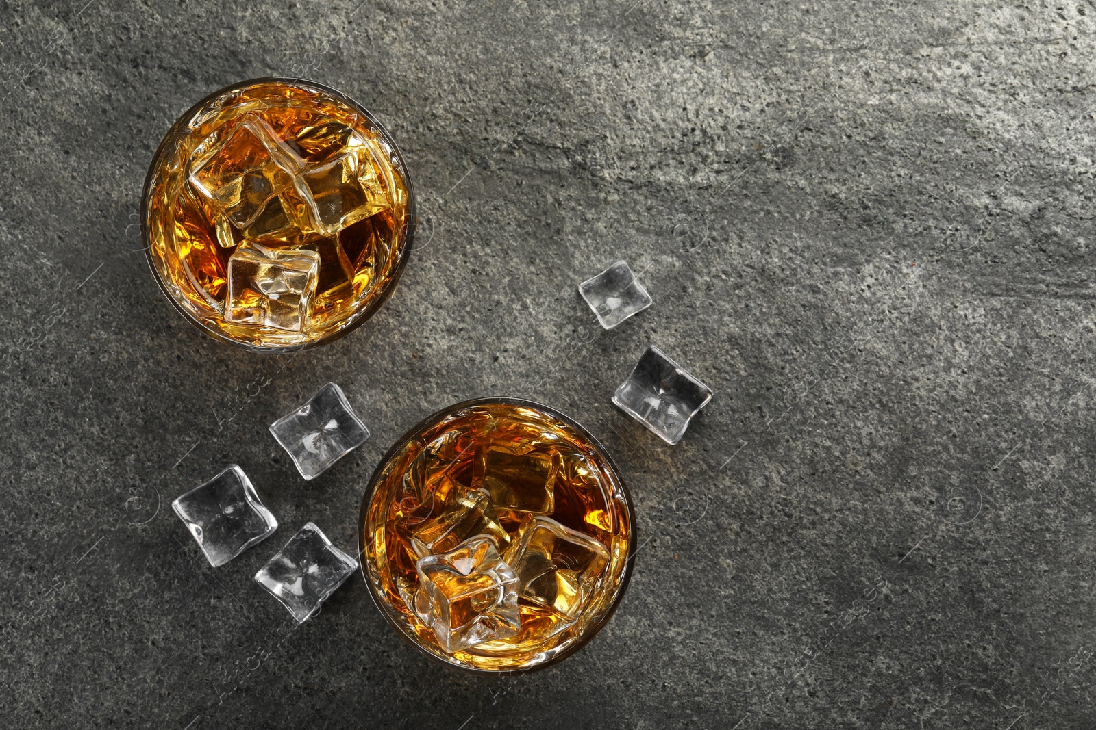 Photo of Whiskey and ice cubes in glasses on grey textured table, top view. Space for text