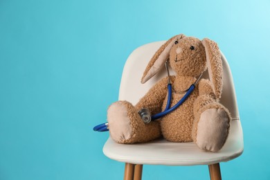 Toy rabbit with stethoscope on chair against light blue background, space for text. Pediatrician practice