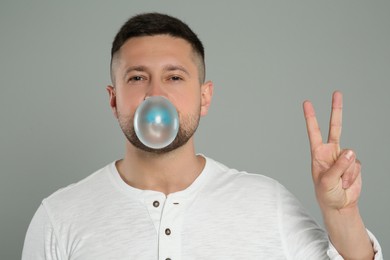 Photo of Handsome man blowing bubble gum and showing peace gesture on light grey background