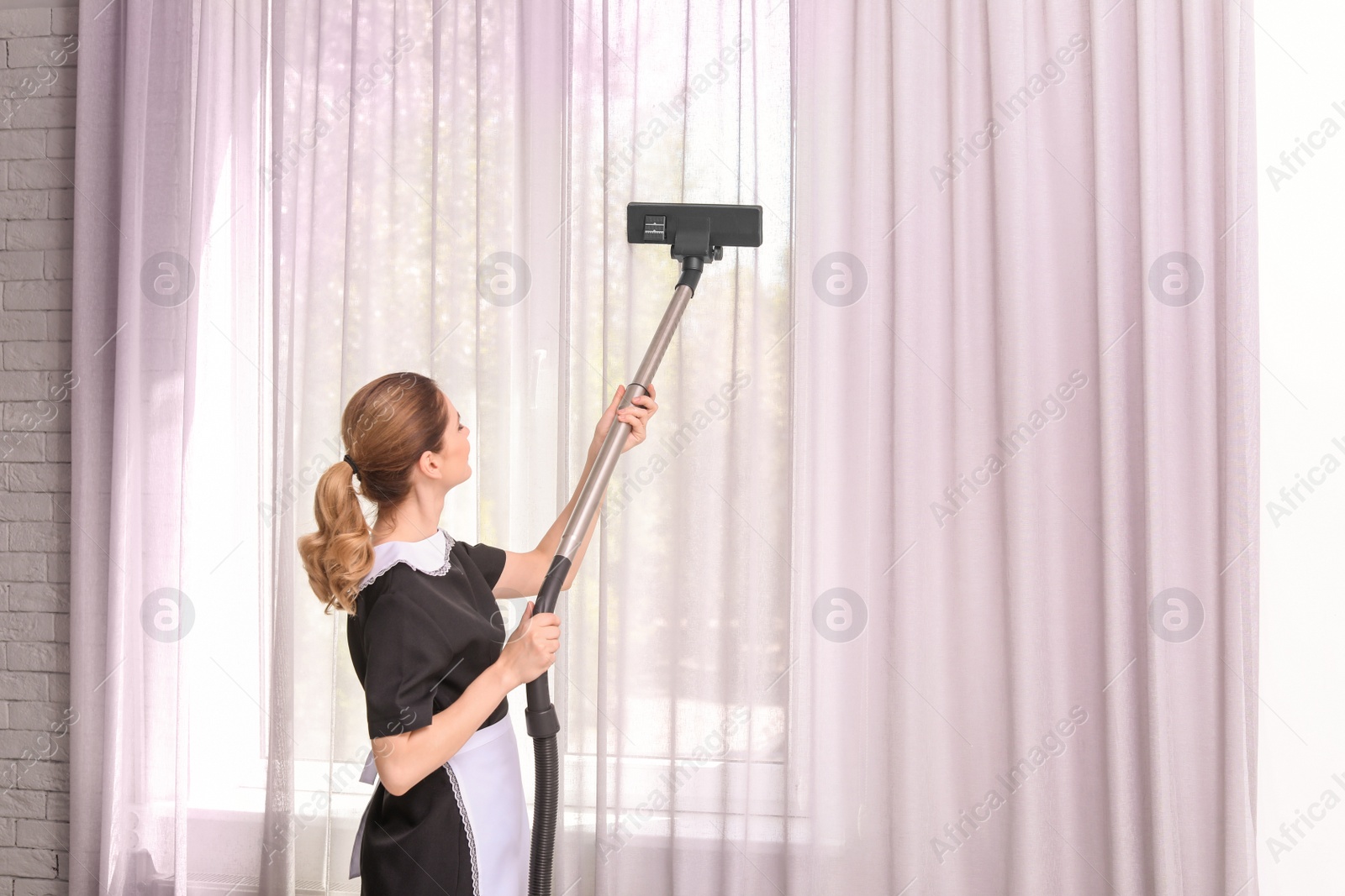 Photo of Female worker removing dust from curtains with professional vacuum cleaner indoors