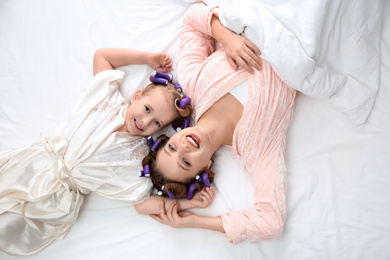 Photo of Happy mother and daughter with curlers lying on bed, top view