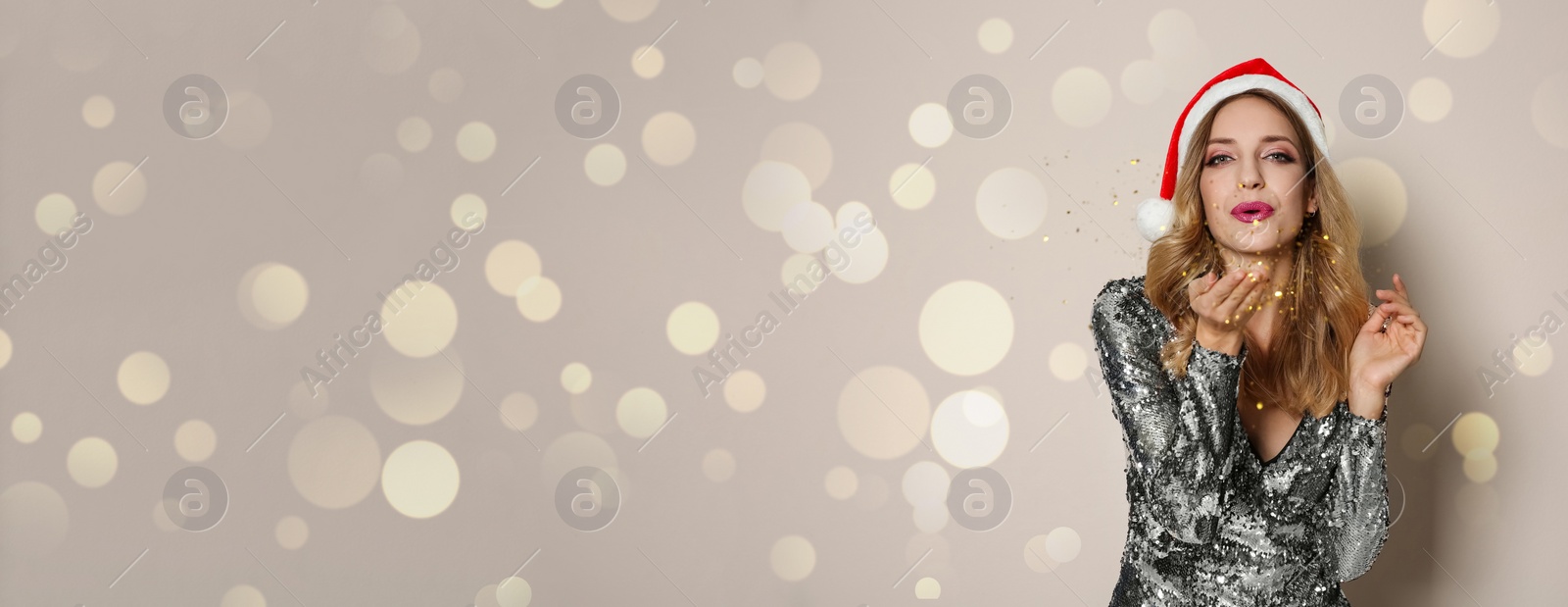 Image of Beautiful woman in Christmas hat blowing away golden glitters on beige background, space for text. Banner design