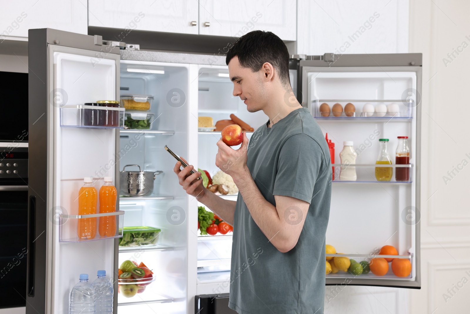 Photo of Happy man with apple and smartphone near refrigerator in kitchen