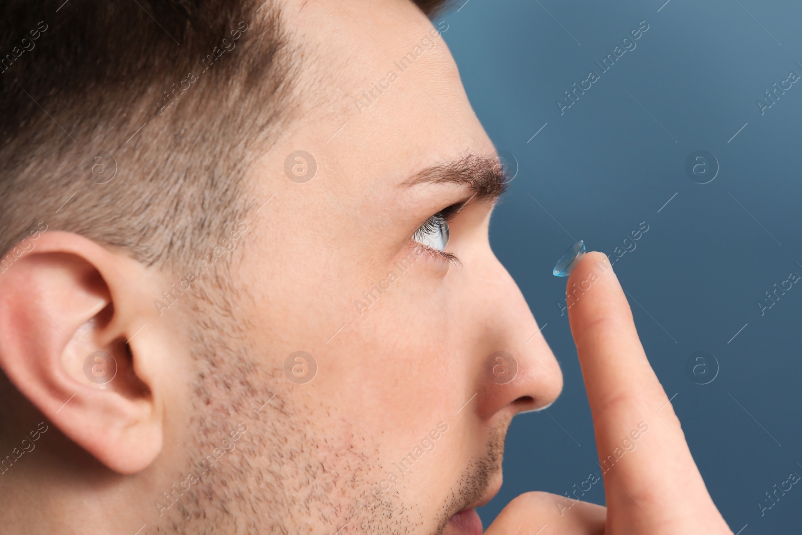 Photo of Young man putting contact lens in his eye on color background