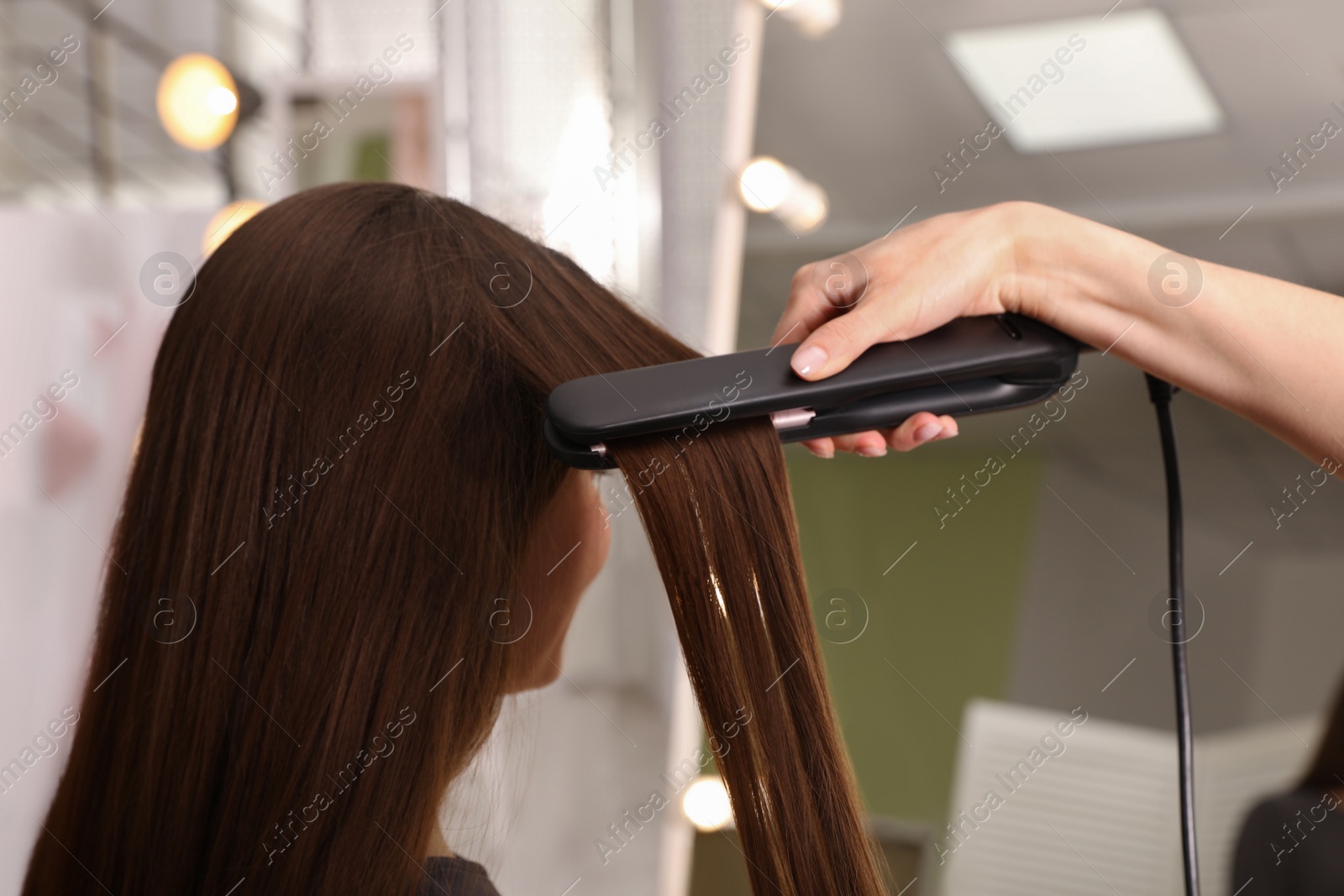 Photo of Stylist straightening woman's hair with flat iron in salon