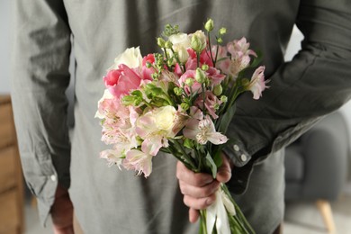 Photo of Man hiding bouquet of flowers for his beloved woman indoors, closeup