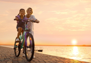 Happy mother teaching her daughter to ride bicycle near river at sunset