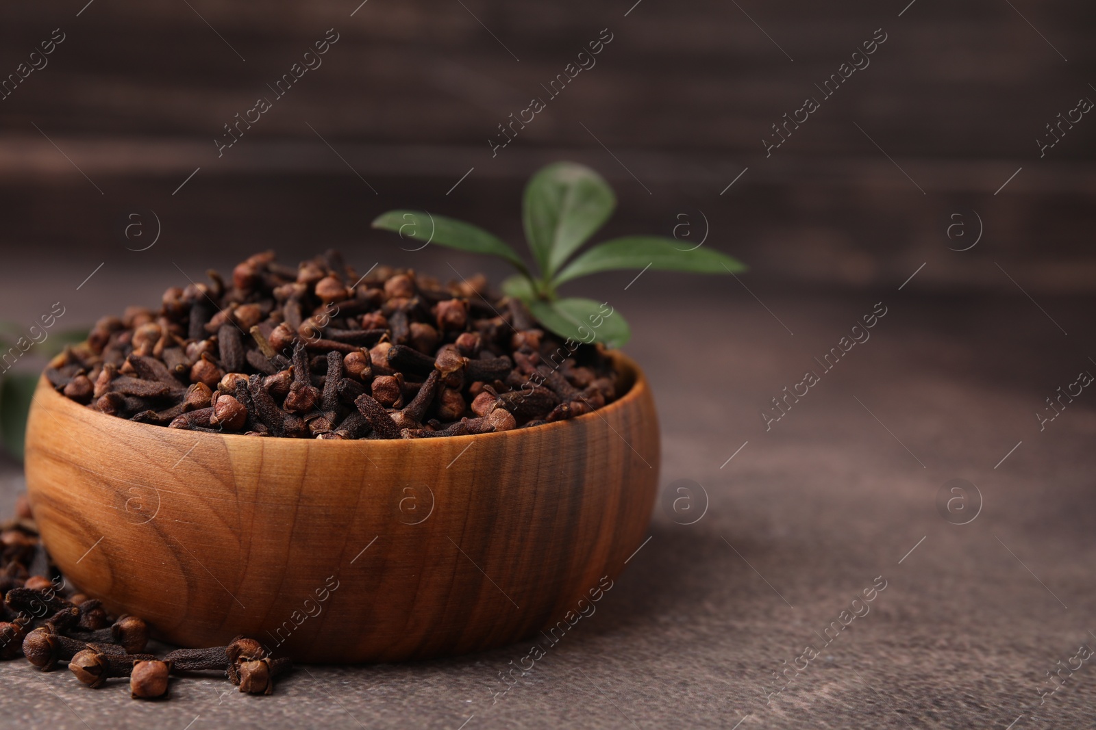 Photo of Aromatic cloves and green leaves in bowl on brown table, closeup. Space for text