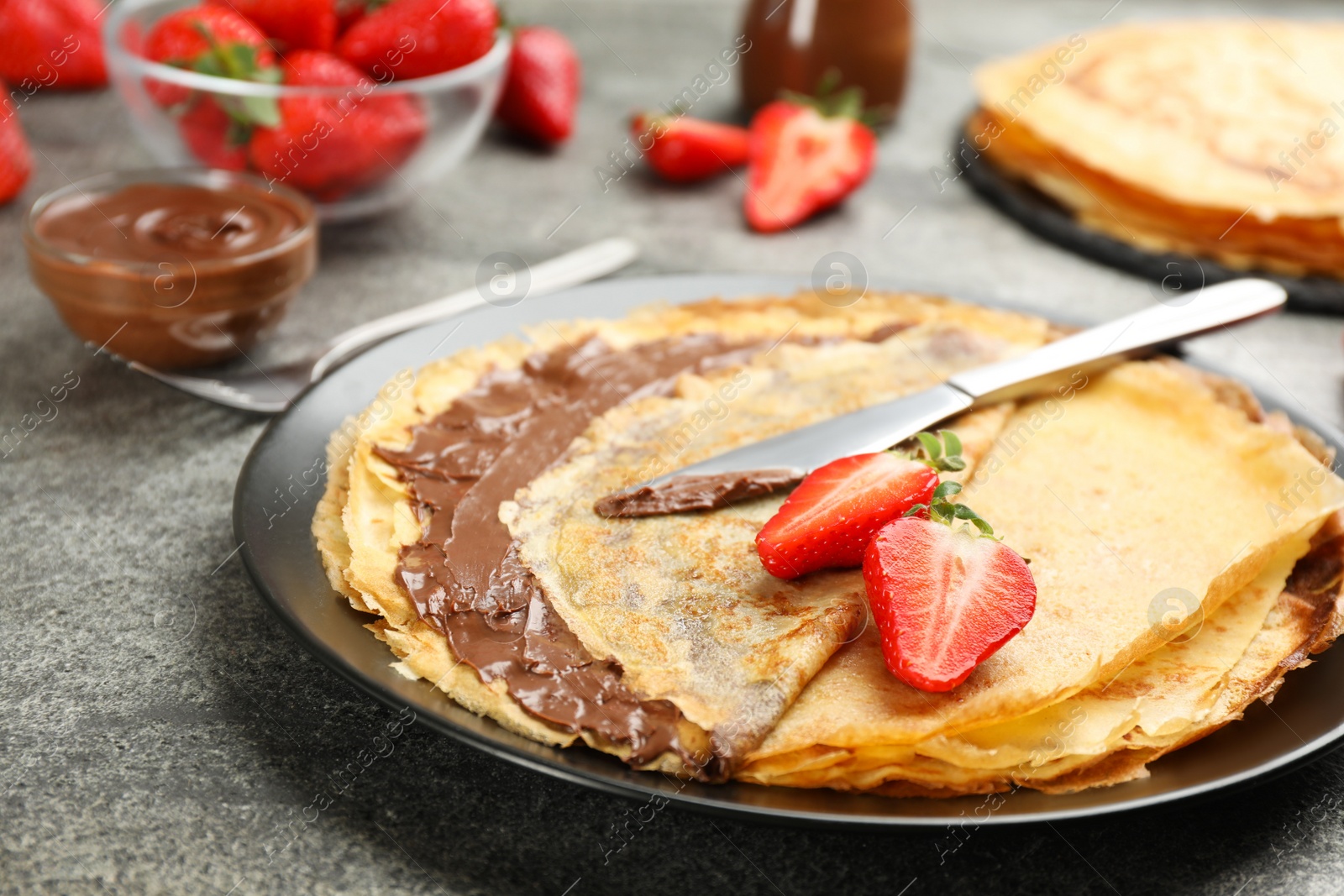 Photo of Delicious thin pancakes with chocolate paste and strawberries on grey table