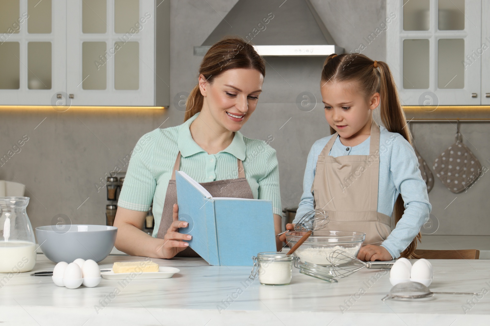 Photo of Cute little girl with her mother cooking by recipe book in kitchen