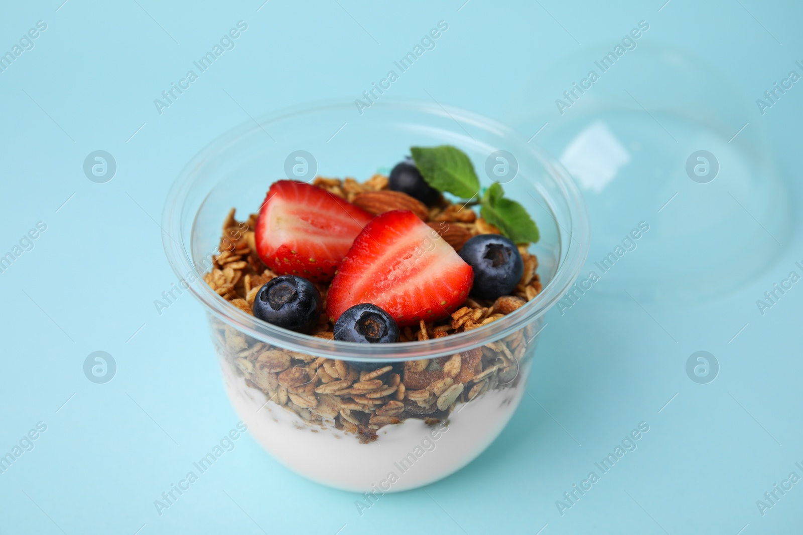 Photo of Tasty granola with berries, nuts and yogurt in plastic cup on light blue background, closeup