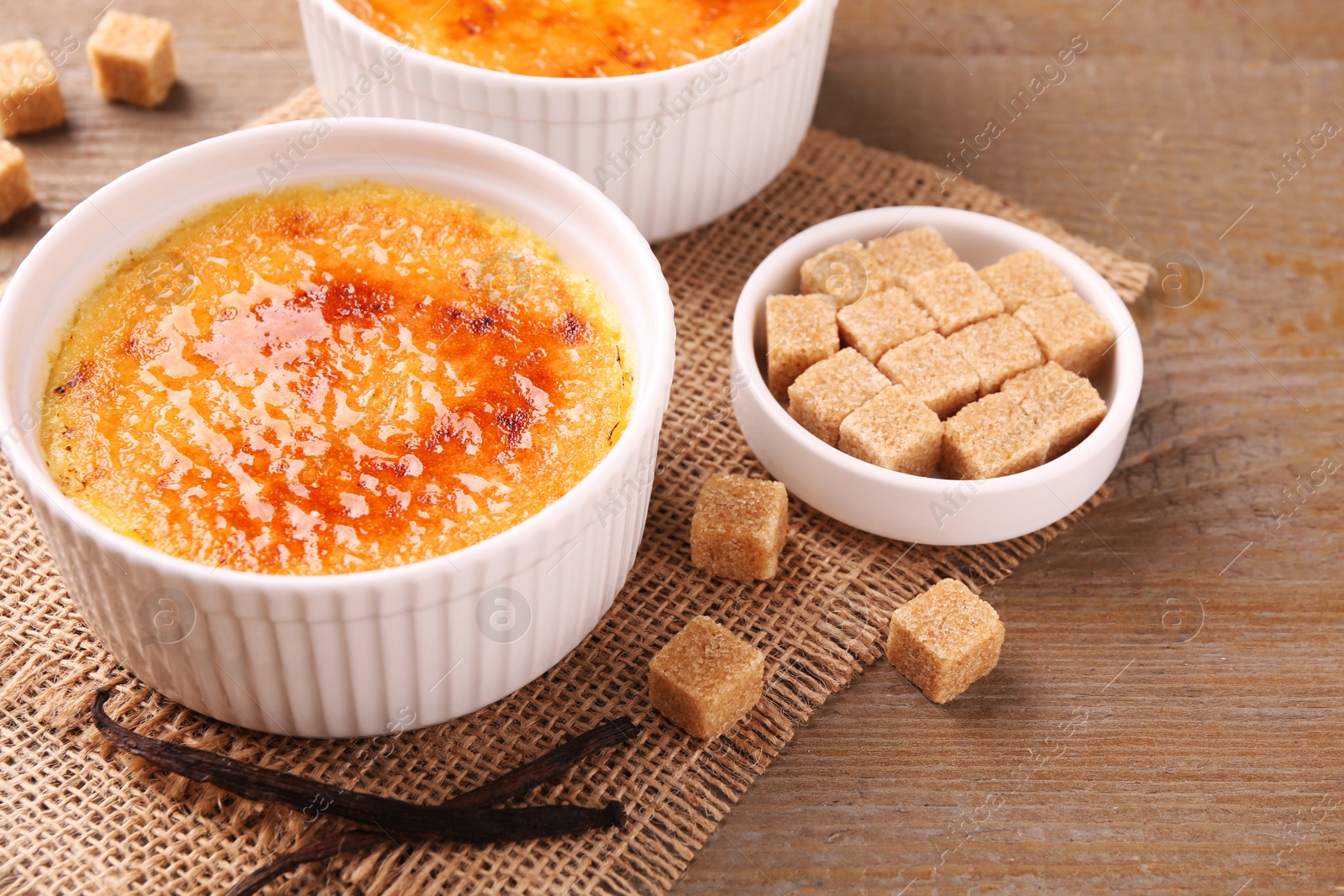 Photo of Delicious creme brulee in bowls, vanilla pods and sugar cubes on wooden table, closeup