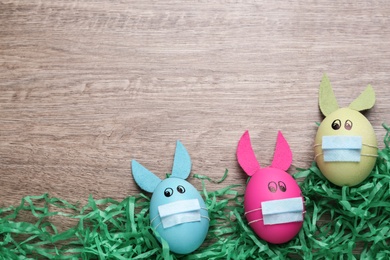 Photo of Dyed eggs with bunny ears in protective masks on wooden background, flat lay and space for text. Easter holiday during COVID-19 quarantine