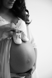 Young pregnant woman in lace nightgown holding baby shoes on light background, black and white effect. Space for text