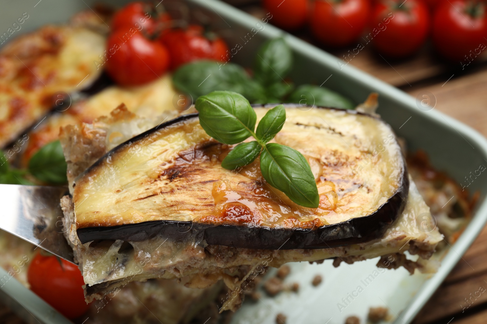 Photo of Spatula with piece of delicious eggplant lasagna over baking dish at table, closeup