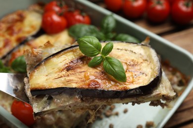 Photo of Spatula with piece of delicious eggplant lasagna over baking dish at table, closeup