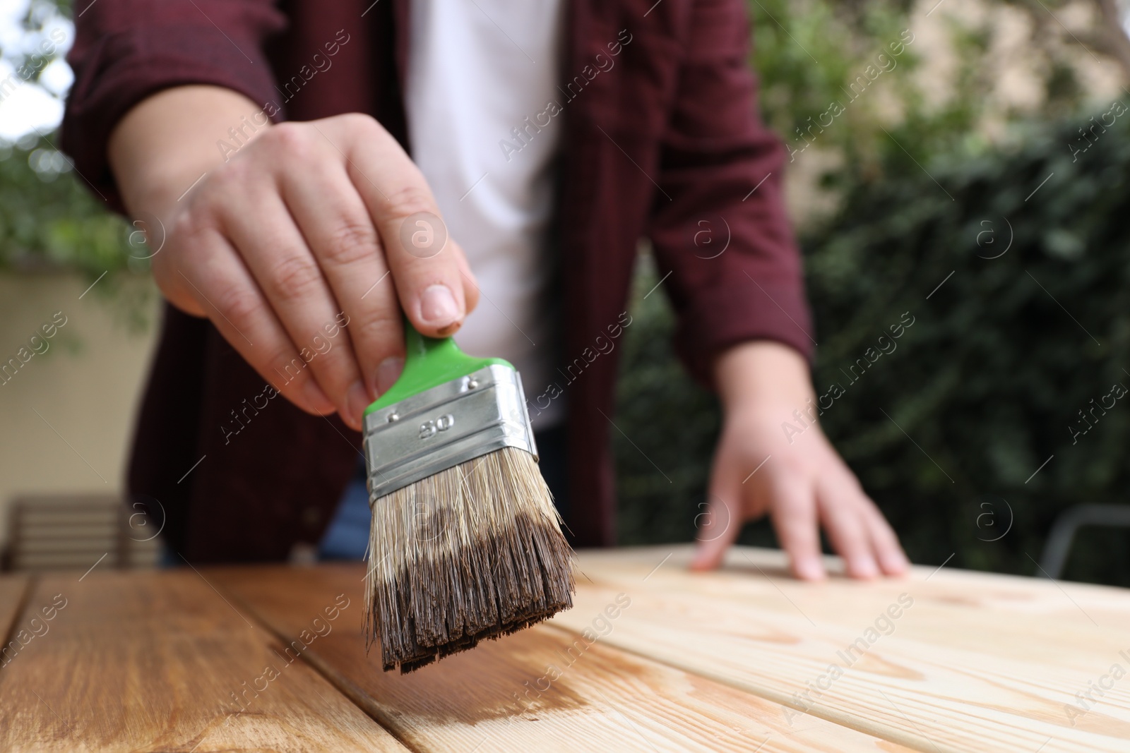 Photo of Man applying wood stain onto wooden surface against blurred background, closeup