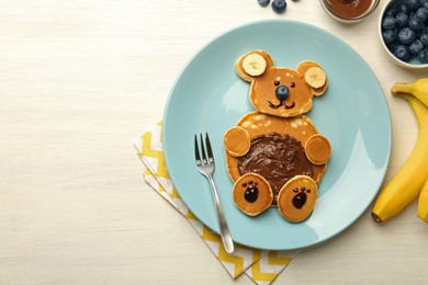 Photo of Creative serving for kids. Plate with cute bear made of pancakes, blueberries, bananas and chocolate paste on light wooden table, flat lay. Space for text