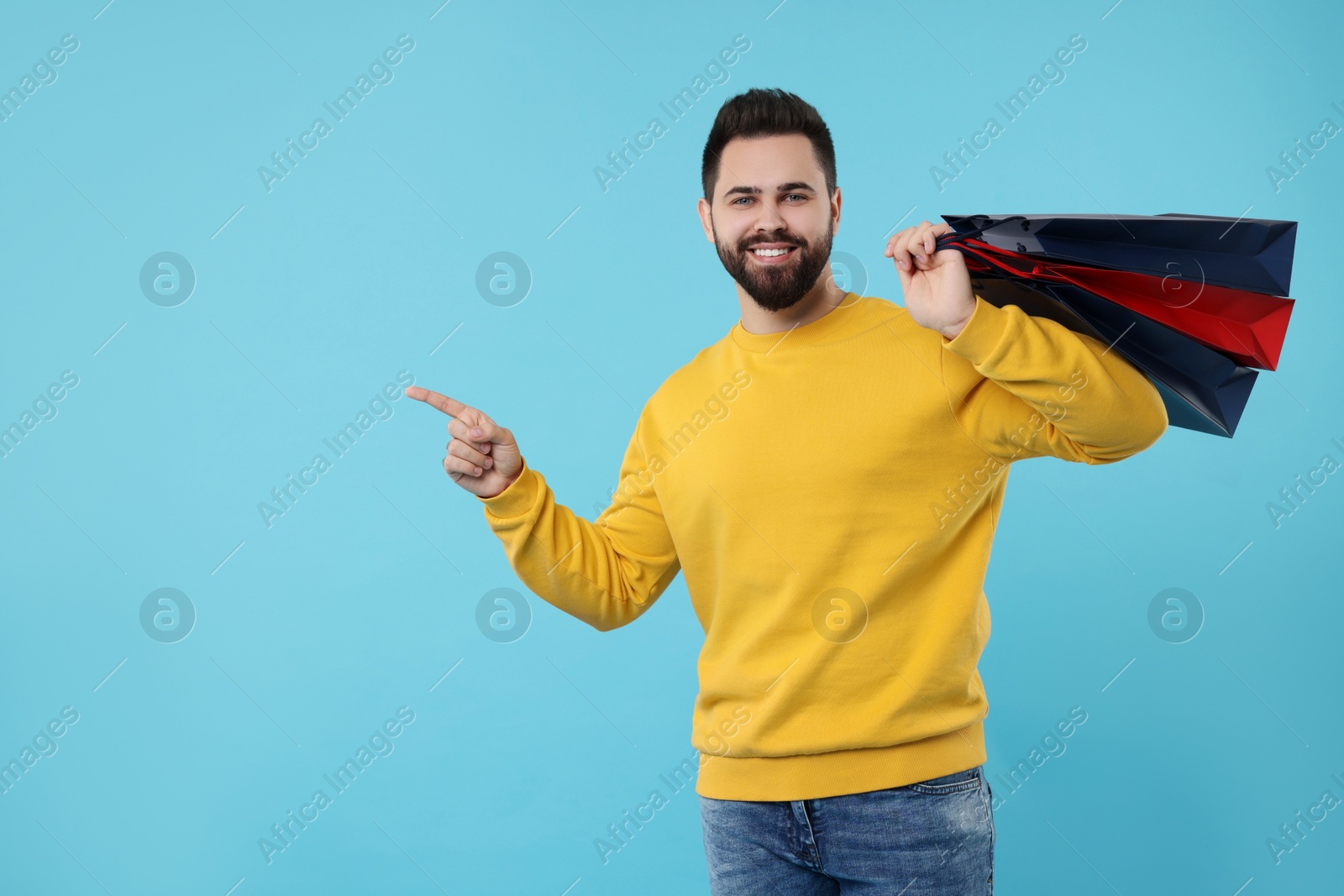 Photo of Smiling man with paper shopping bags pointing at something on light blue background. Space for text