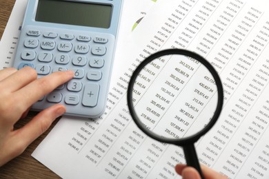 Photo of Woman looking at accounting document through magnifying glass while using calculator at table, closeup