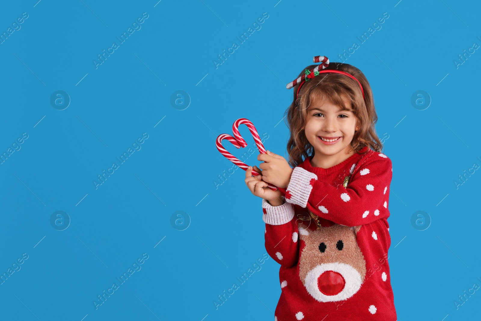 Photo of Cute little girl in Christmas sweater making heart shape with candy canes against blue background. Space for text