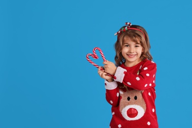 Cute little girl in Christmas sweater making heart shape with candy canes against blue background. Space for text