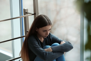 Photo of Upset teenage girl sitting at window indoors