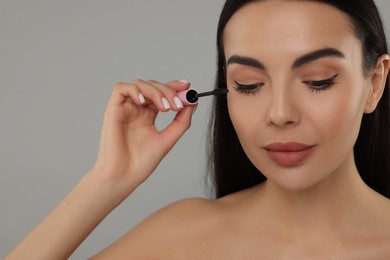 Beautiful young woman applying mascara on grey background