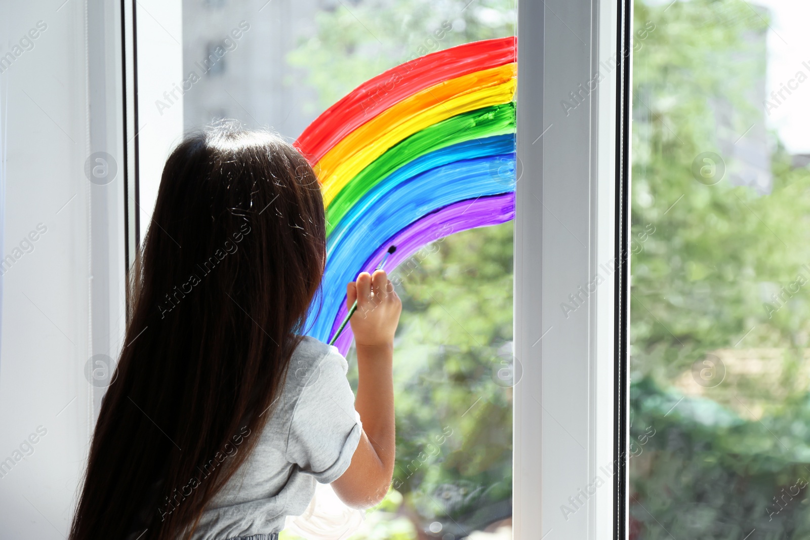 Photo of Little girl drawing rainbow on window indoors. Stay at home concept