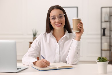 Photo of Young woman with cup of coffee writing in notebook at white table indoors