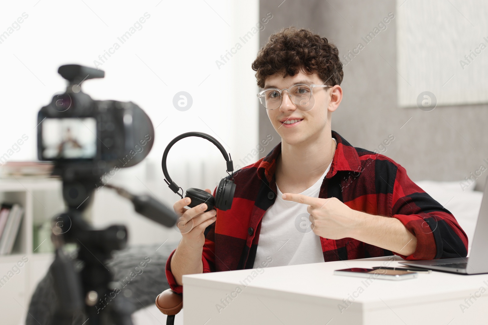 Photo of Smiling teenage blogger pointing at his headphones while streaming at home