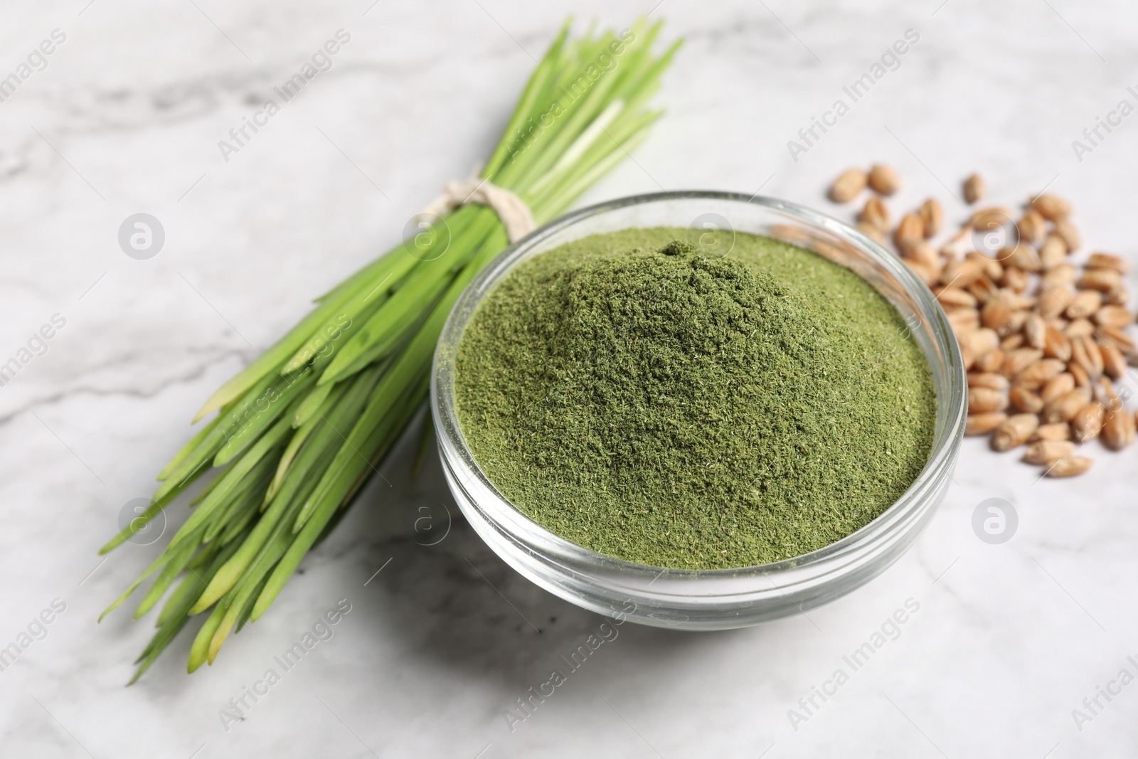 Photo of Wheat grass powder in glass bowl, seeds and fresh sprouts on white marble table, closeup